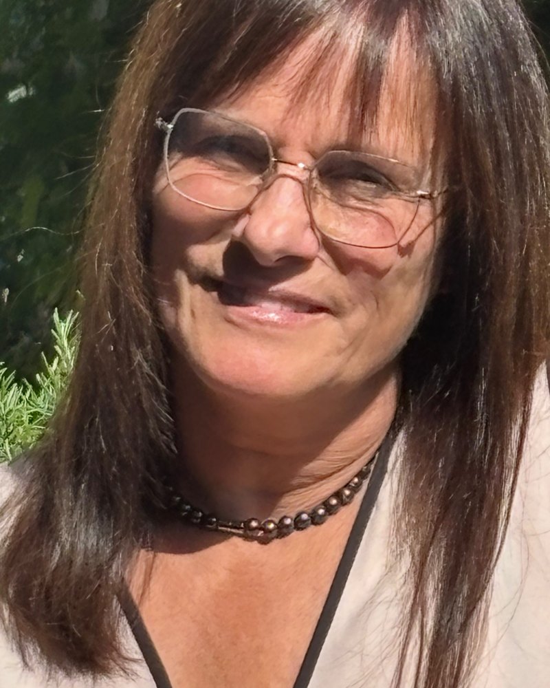 Porträt von Kerstin Siegismund: lächelnde Frau mit Brille und langen braunen Haaren vor grünem Laub.